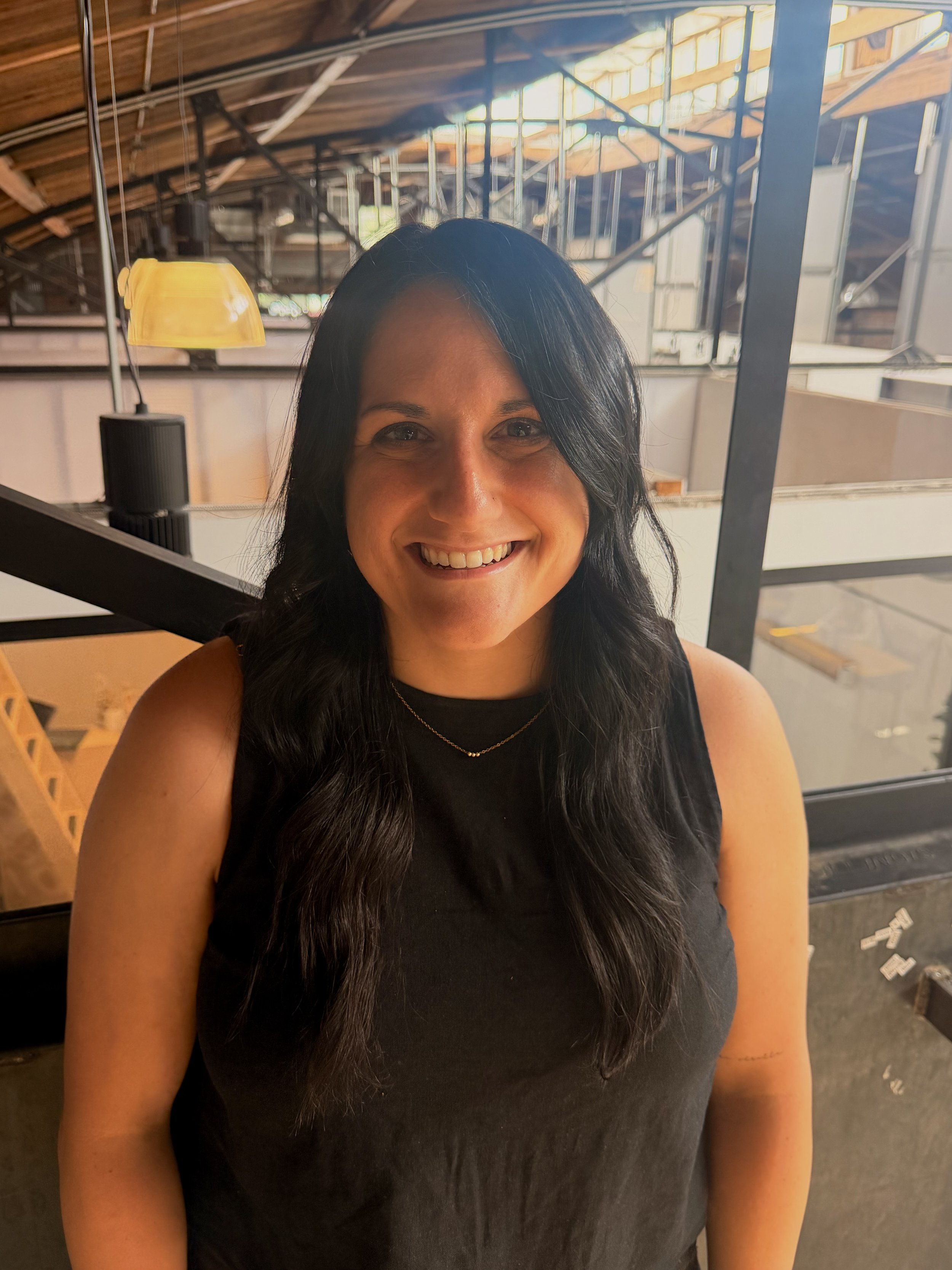 Devan Grayson at Sunergos Counseling Collective smiling, wearing a beige sweater, with a background of a modern indoor space with visible beams and large windows.
