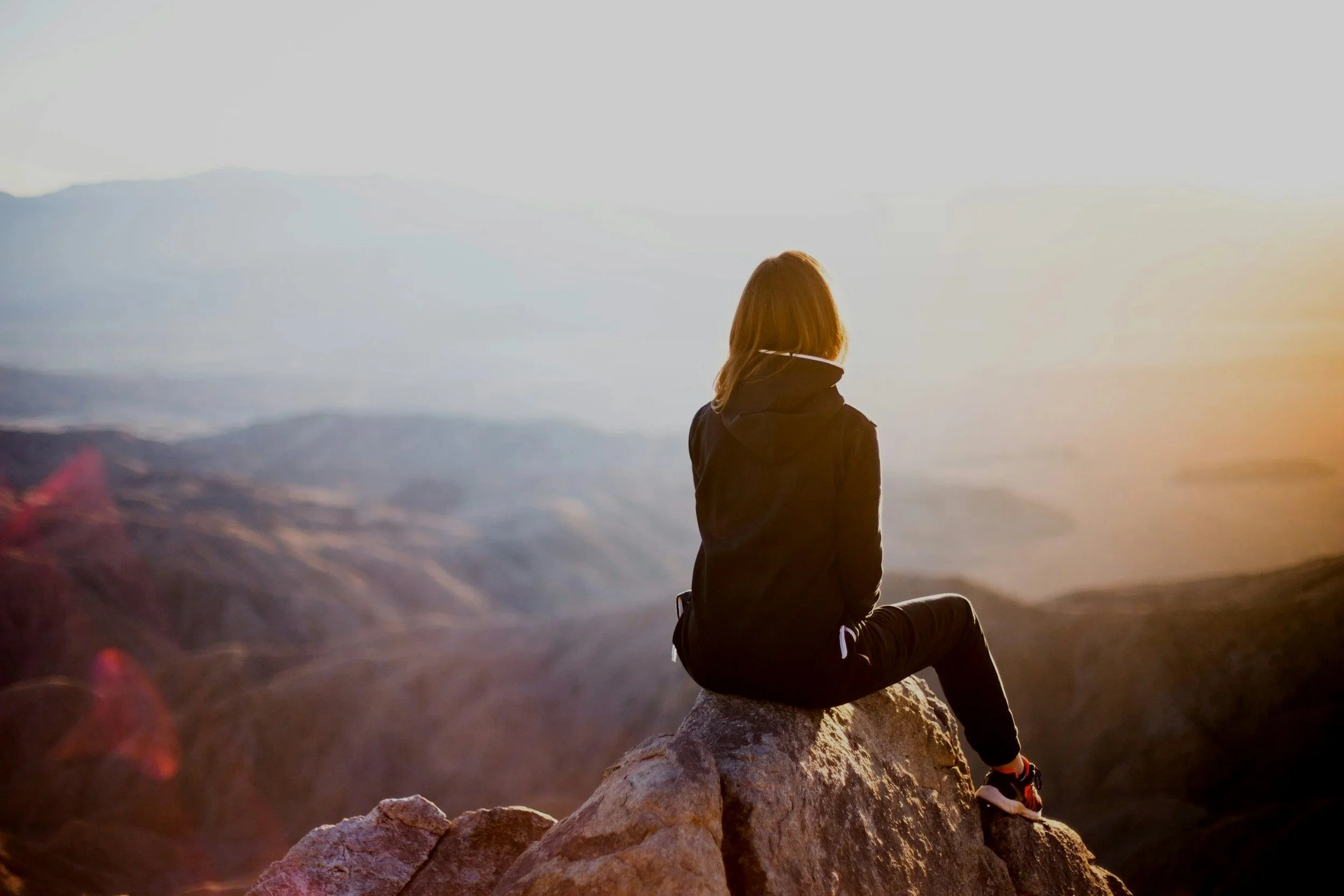 Person sitting on rocks, facing mountains at sunset or sunrise.