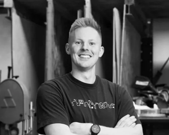 A smiling young man standing in a workshop or garage, arms crossed, wearing a T-shirt and a watch.