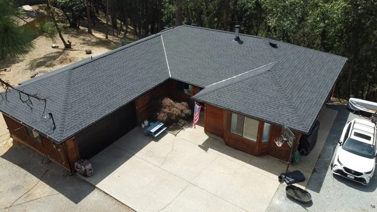 Aerial view of a single-story house with a gray roof, surrounded by trees. The driveway has a parked car, and there's an American flag displayed by the entrance.