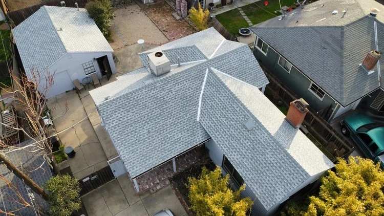Aerial view of residential houses with gray shingled roofs, driveways, and yards. Two houses and a detached garage are visible with a driveway and parked cars. Small trees and other vegetation are present in the yards.