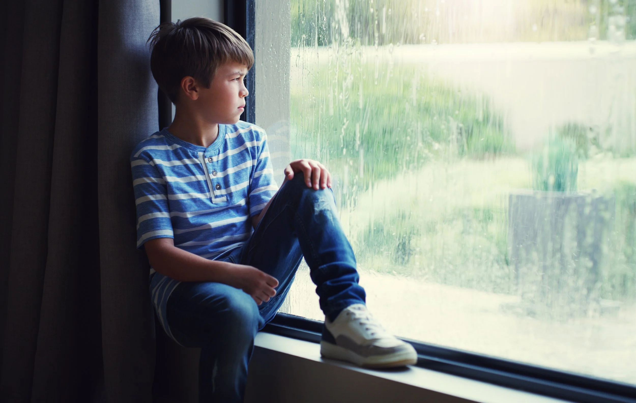 Boy sitting on a windowsill, looking outside at rainy weather.