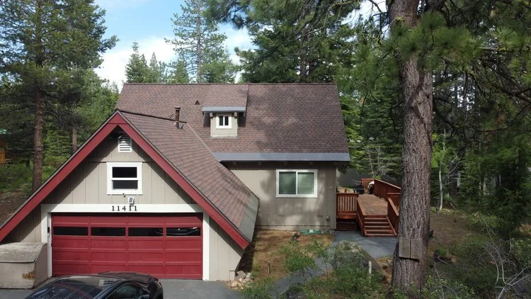 A-frame house with a garage, surrounded by pine trees, featuring a brown shingled roof, red garage door, and wooden deck. Located in a forested area.