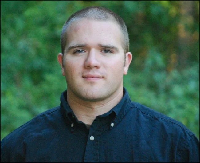 Young man with short hair wearing a navy blue button-up shirt outdoors with green foliage in the background.