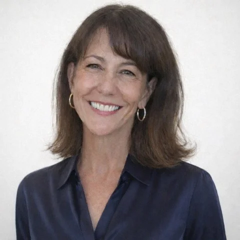 Portrait of a woman with shoulder-length brown hair, smiling, wearing a navy blue collared shirt, and gold hoop earrings, standing against a plain white background.
