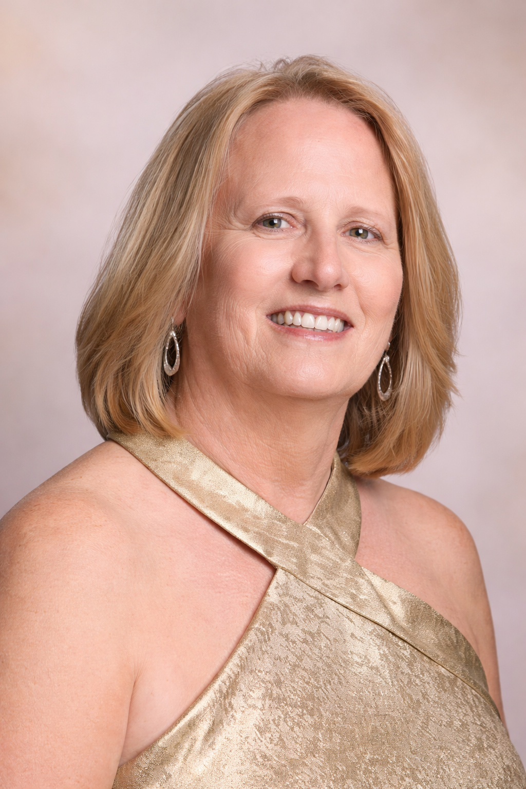 A woman with shoulder-length blonde hair smiling, wearing a gold halter dress and dangling earrings.