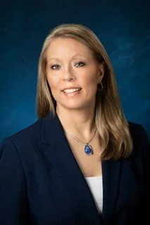 Professional portrait of a woman with long, light brown hair wearing a dark blazer, white blouse, and a blue pendant necklace against a blue background.