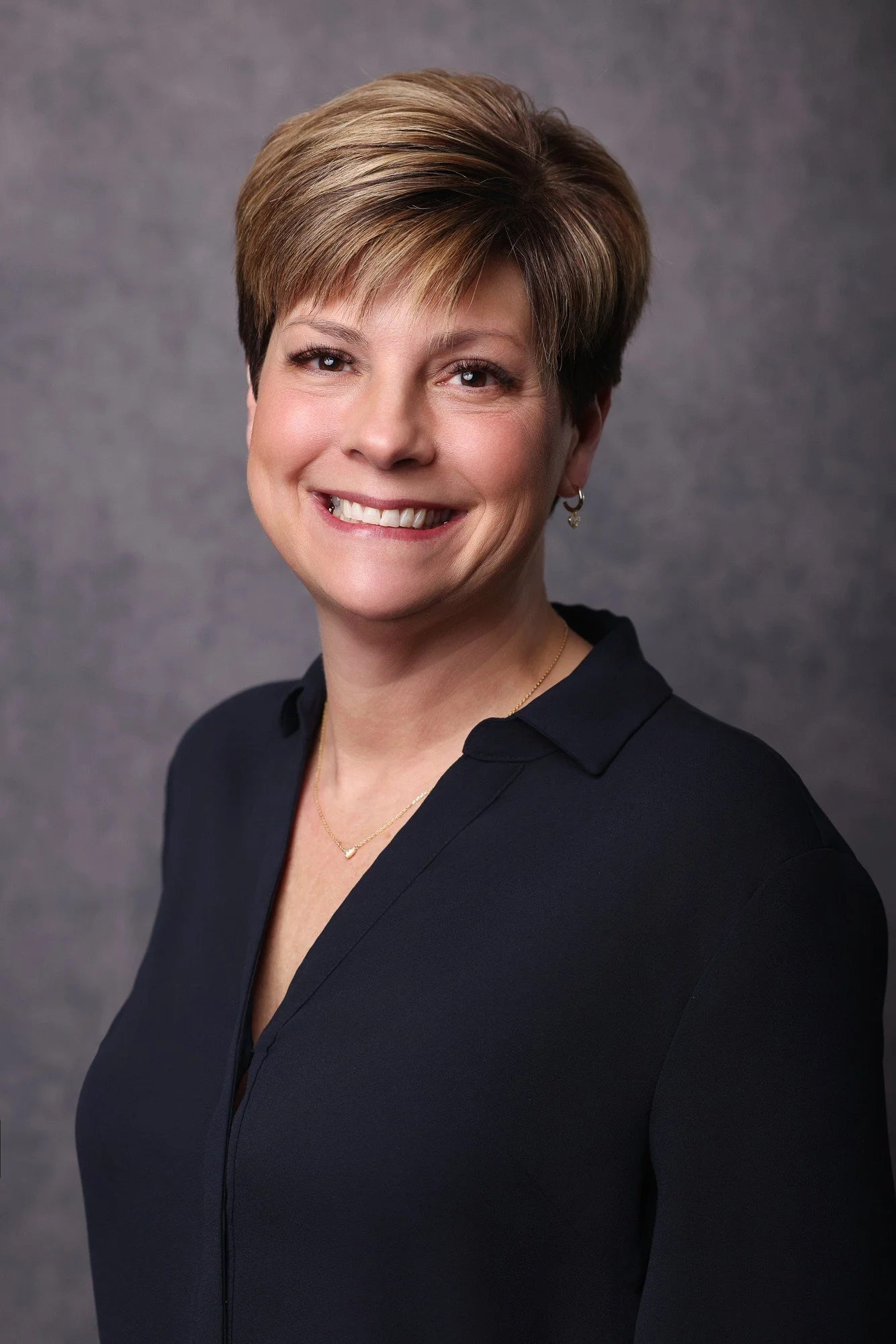 A smiling woman with short, styled brown hair, wearing a black collared top, gold jewelry, earrings, and a necklace, against a neutral gray background.