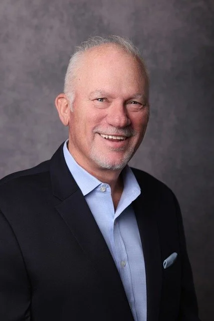 A smiling middle-aged man with light-colored hair and a beard, wearing a dark blazer over a light blue button-up shirt, standing against a gray background.