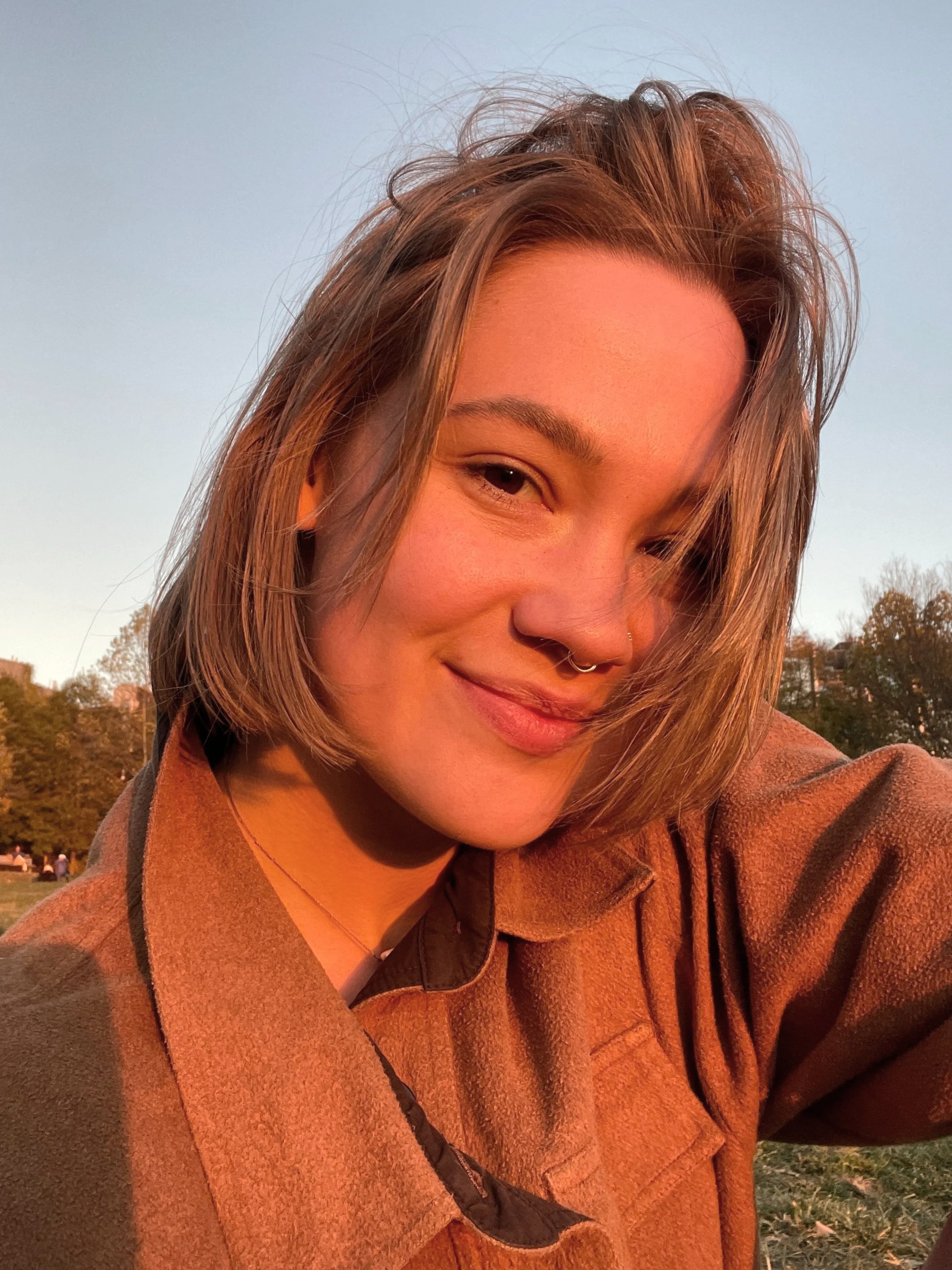 A young woman with light skin and short brown hair smiling during sunset outdoors.