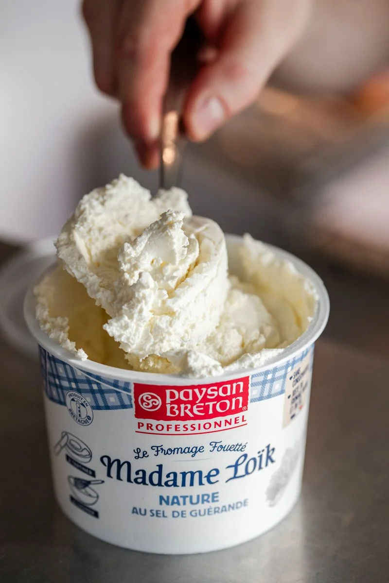 Une main utilise un couteau pour couper de la crème de fromage dans un pot de Madame Loïk, un fromage fouetté au sel de Guérande.
