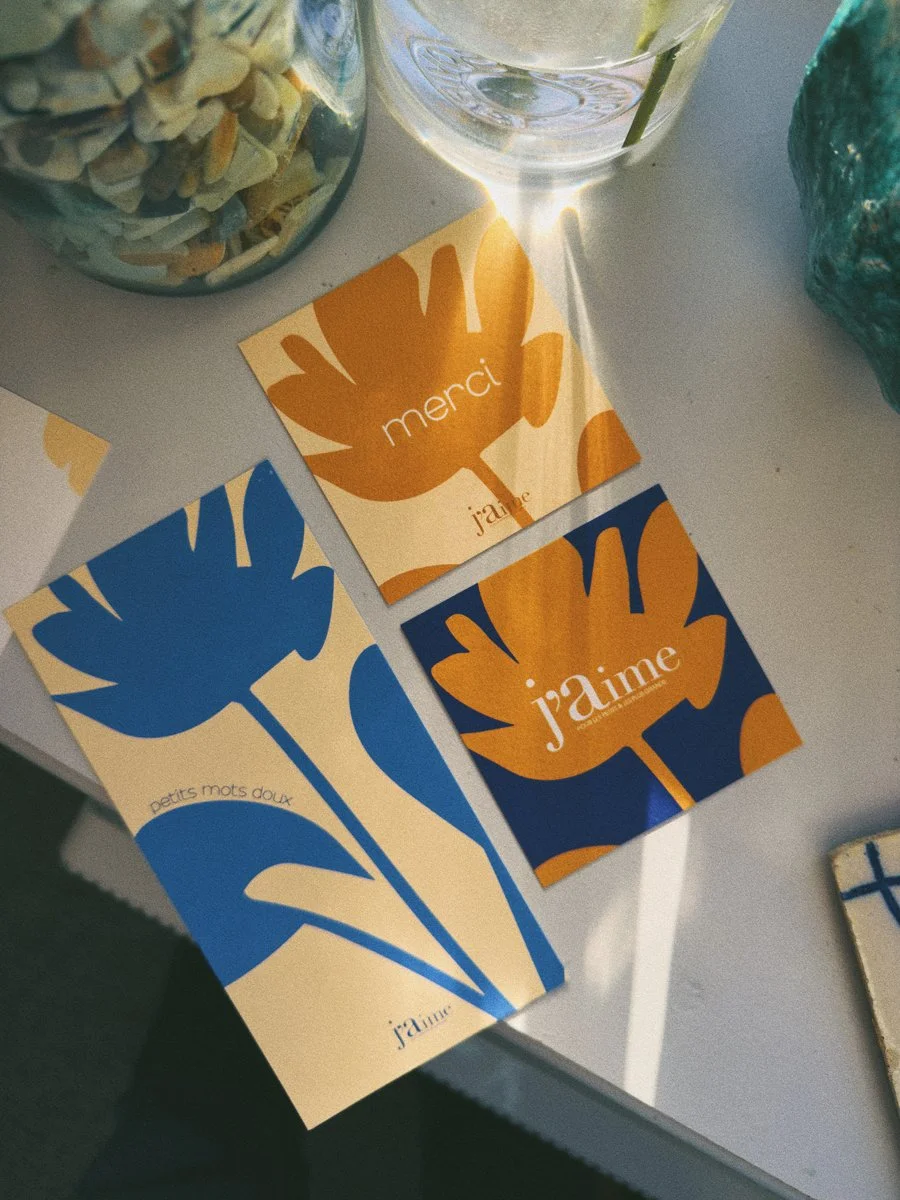 Cartes avec des motifs floraux en bleu et orange, contenant les mots "merci" et "j'aime" sur une table, près d'un verre d'eau et des coquillages dans un bocal.