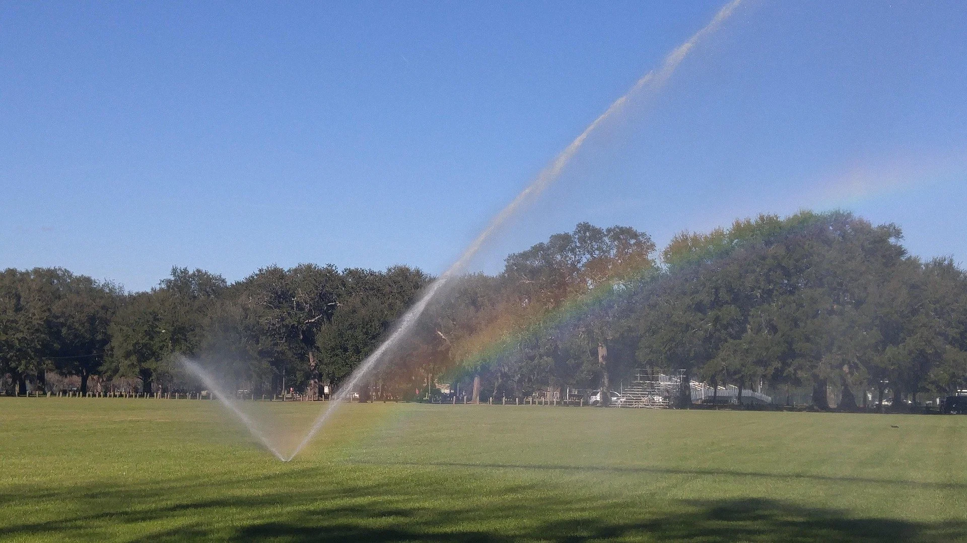 A park with a sprinkler creating water spray, forming a faint rainbow in a clear blue sky with trees in the background.
