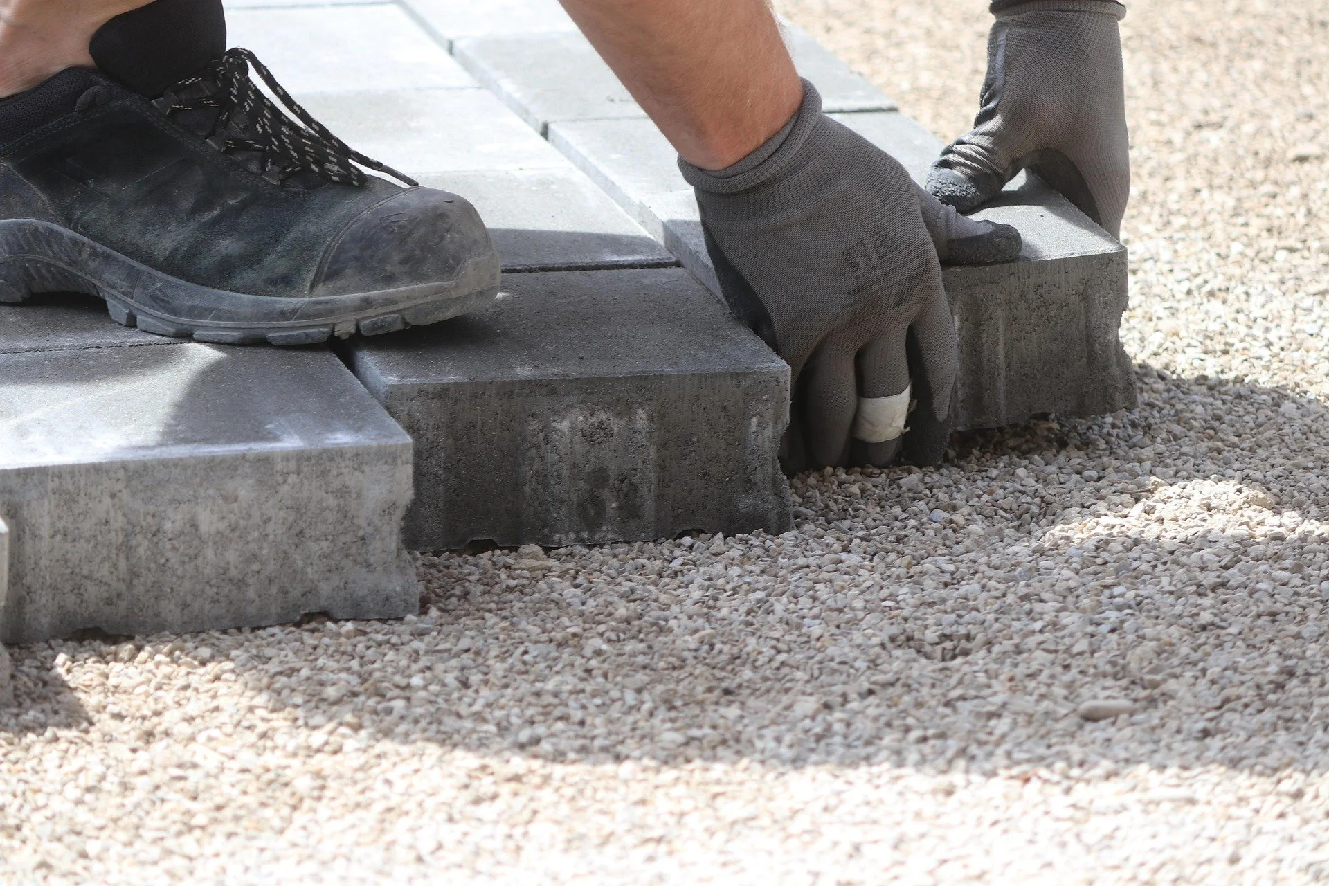 A worker placing concrete bricks on a gravel ground while wearing gloves and black work shoes.