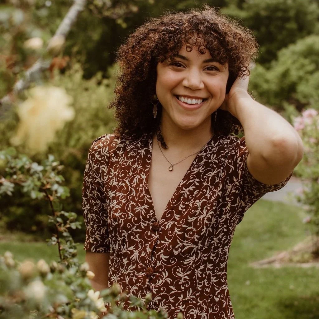 A person with curly hair smiling, wearing a patterned brown dress, standing in a garden with greenery and flowers.