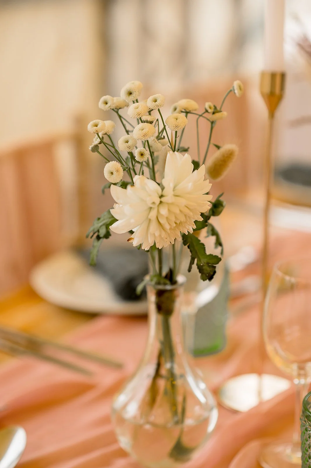 A small bouquet of white flowers in a clear glass vase on a pink tablecloth with dinnerware and wine glasses in the background.