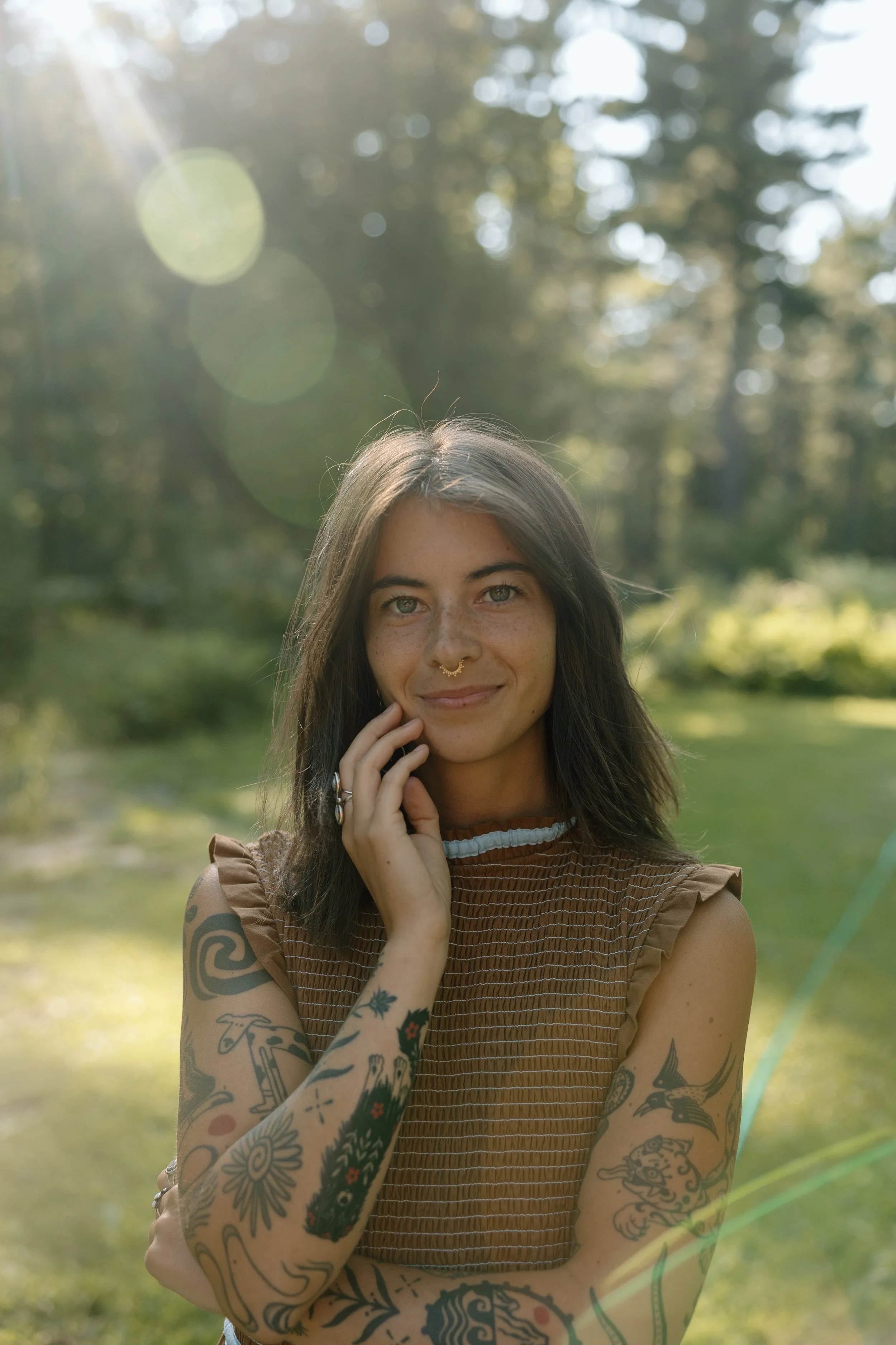 A young woman with dark hair and tattoos on her arms, smiling, outdoors with sunlight and trees in the background.