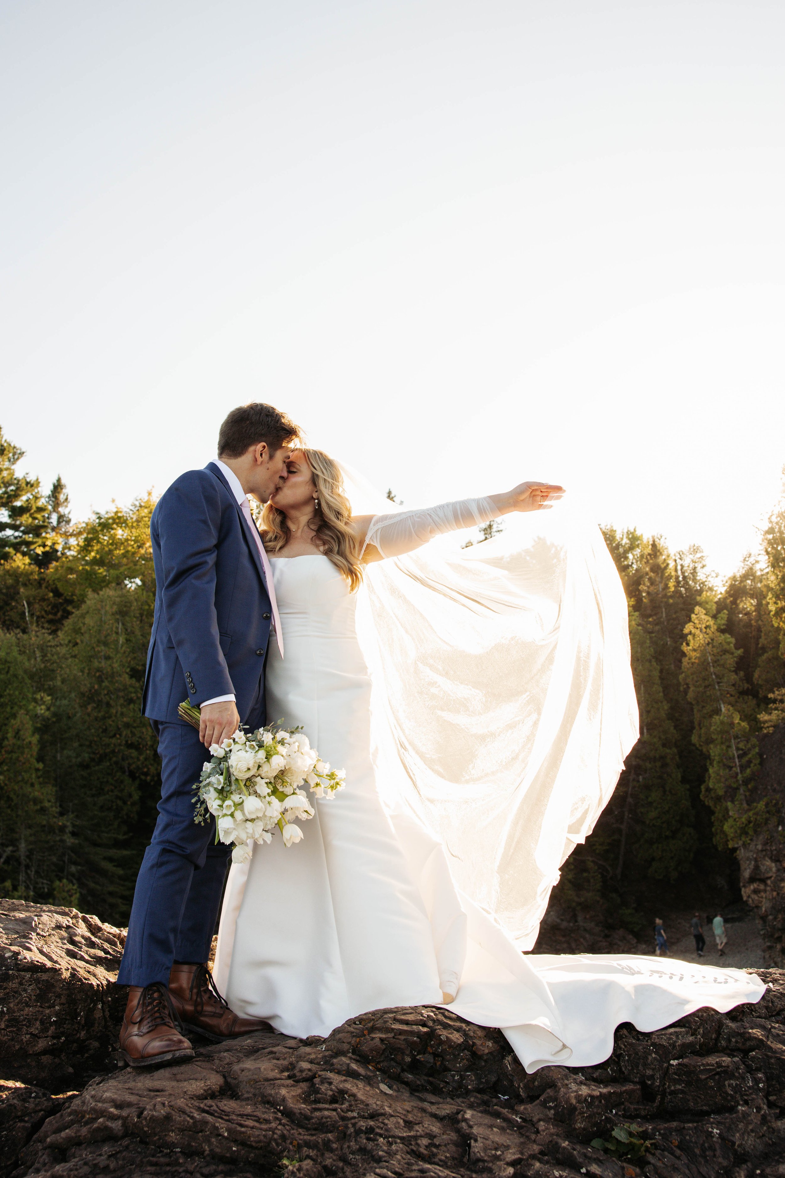 A bride and groom kissing on rocks during a sunset outdoor wedding, with the bride holding a bouquet of white flowers and the groom wearing a blue suit.