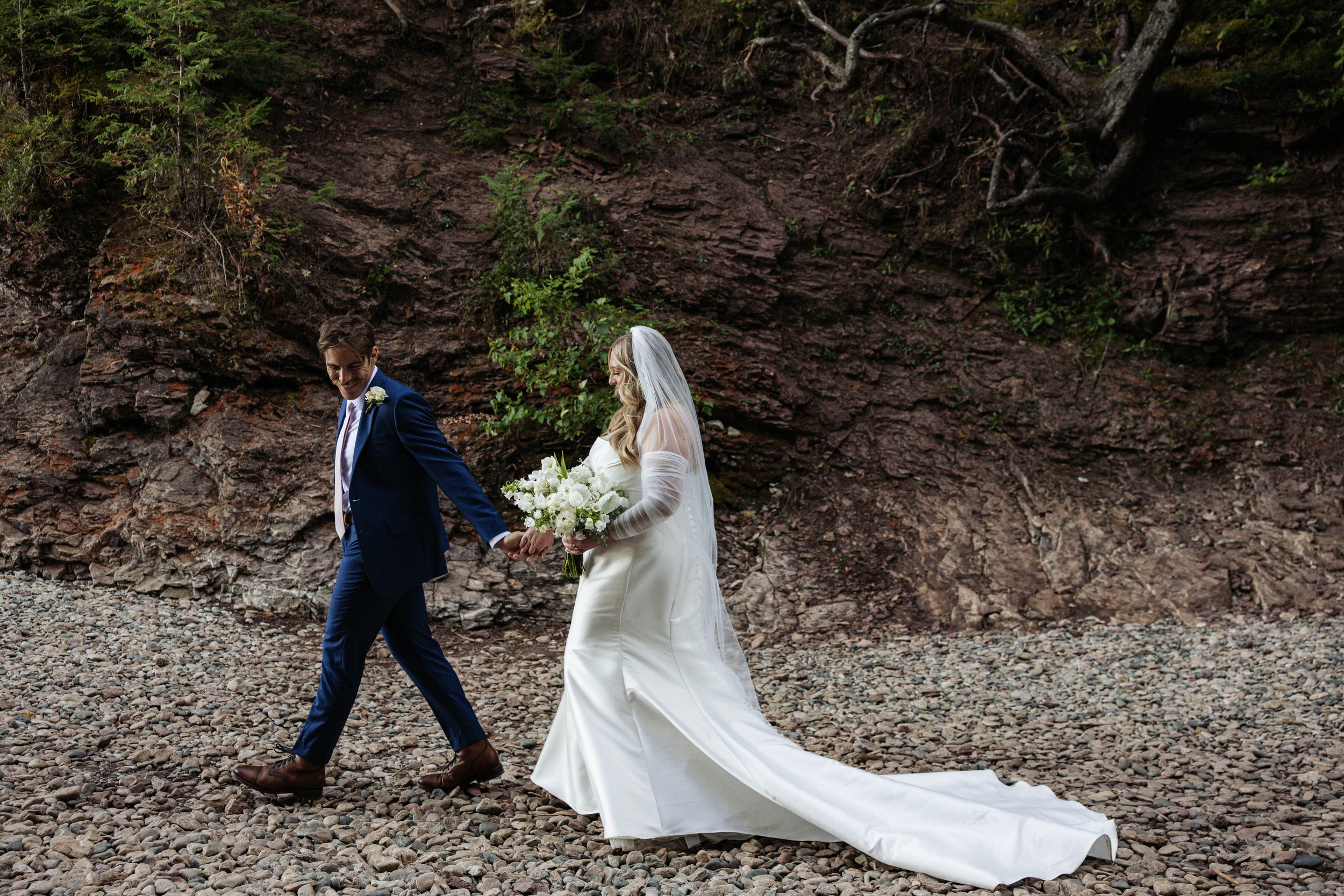 A bride and groom holding hands and walking on a rocky beach. The bride is wearing a white wedding dress with a train and veil, holding a bouquet of flowers. The groom is wearing a dark blue suit with brown shoes, smiling.