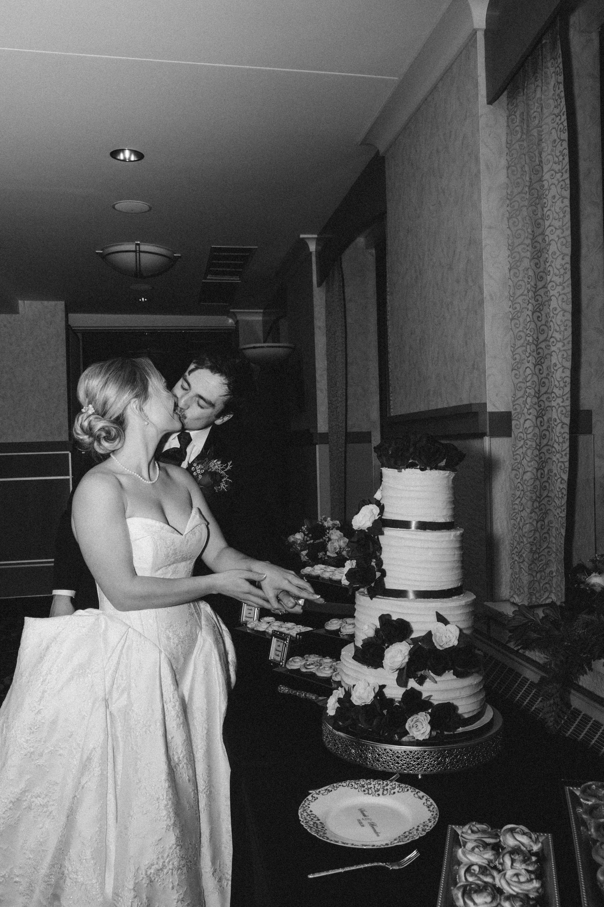 A bride and groom kissing while cutting a wedding cake at a reception hall.
