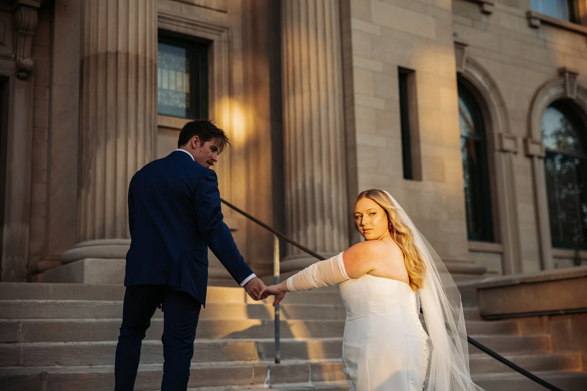 A bride in a white wedding dress and veil holding hands with a groom in a blue suit on the steps of a stone building at sunset.