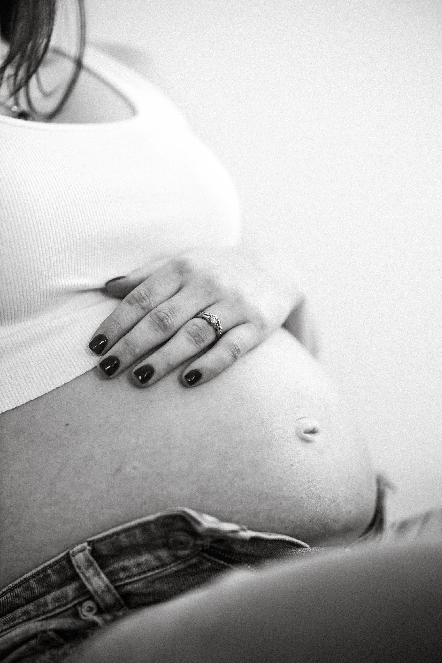 Close-up of a woman's pregnant belly, with her hand resting on it, wearing a ring, black nail polish, and a white top, denim shorts, and a smooth background.