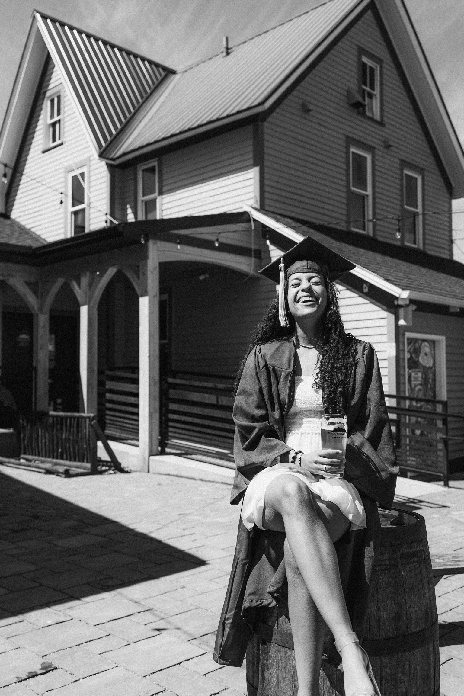 A young woman in a graduation cap and gown smiling and holding a drink while sitting on a barrel outside a house.