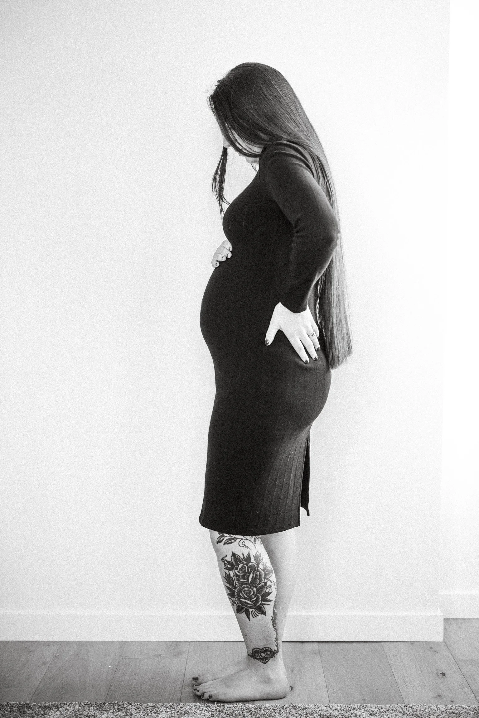 A woman standing barefoot on a wooden floor, wearing a black dress, with tattoos on her lower leg, looking downward with her hands on her hips against a plain wall.