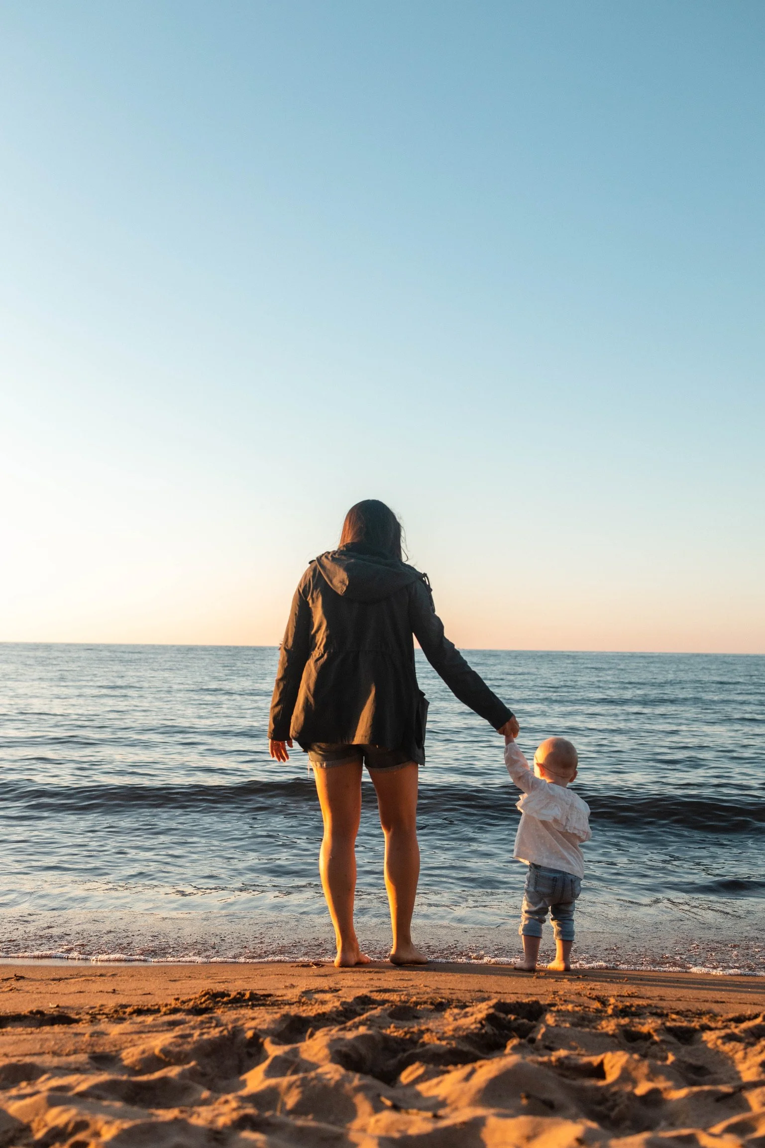 Woman and a young child standing on the beach near the water during sunset, holding hands.
