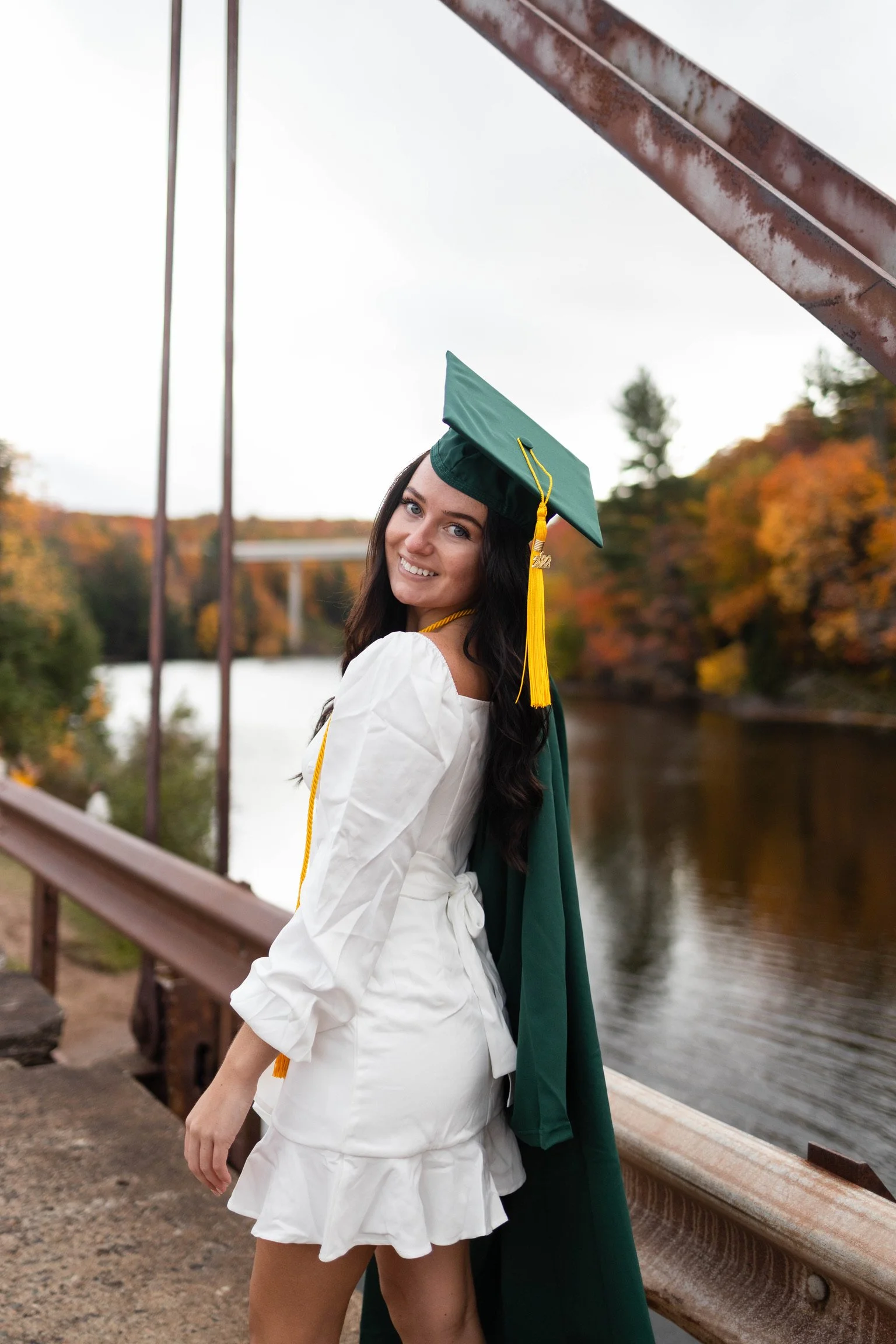 A young woman in a white dress and green graduation cap and gown, smiling outdoors by a river during fall, with colorful autumn trees and a bridge in the background.