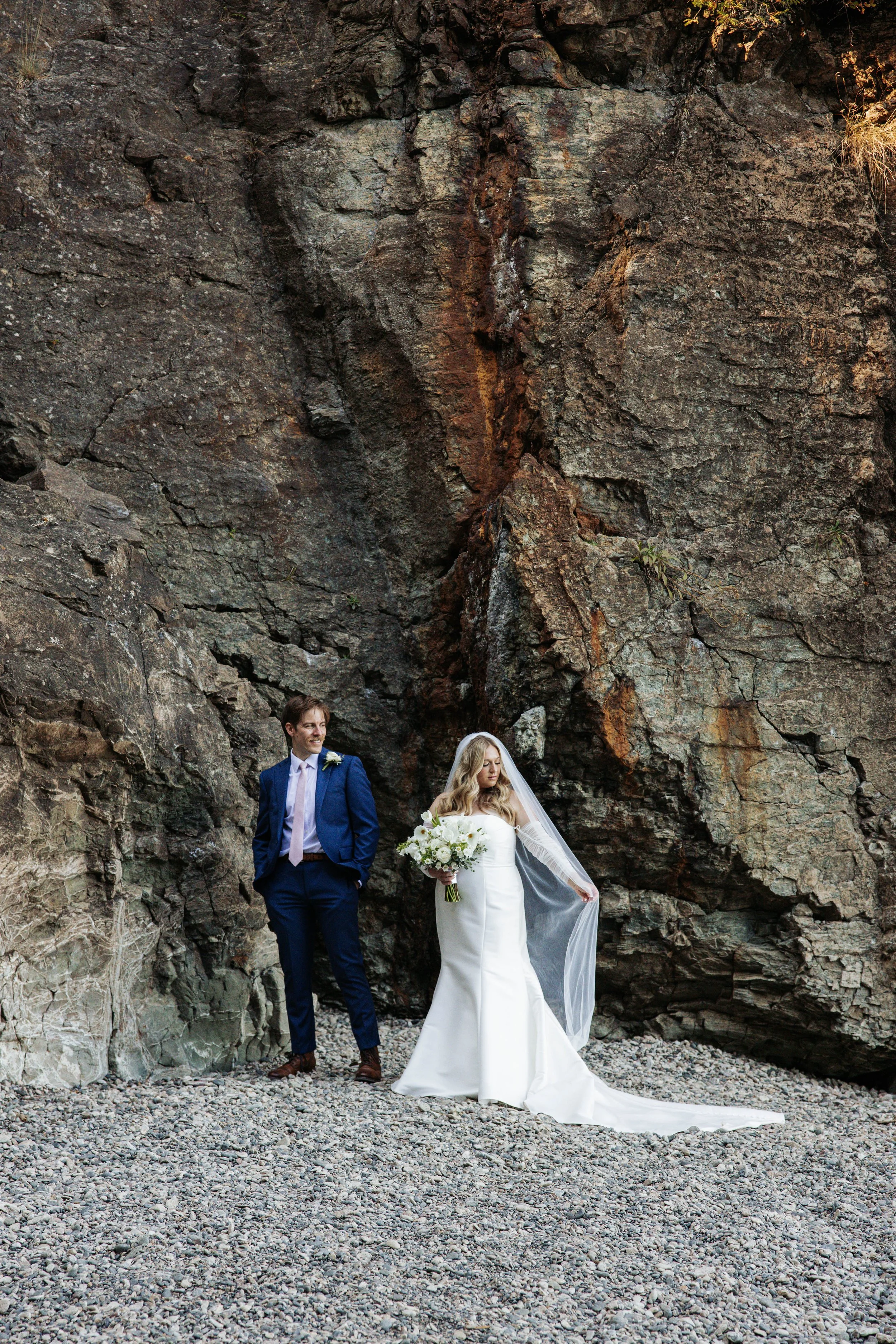 Bride in a white wedding dress holding a bouquet of white flowers, standing near a groom in a blue suit against a rugged stone cliff.