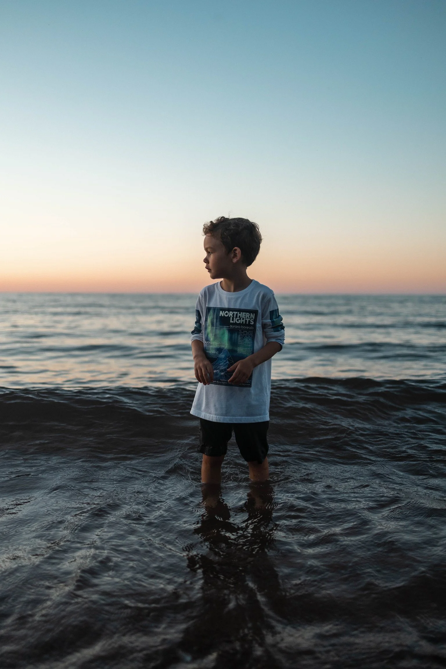 A young boy standing in shallow water at the beach during sunset, wearing a white long-sleeve shirt and dark shorts, looking to the side.