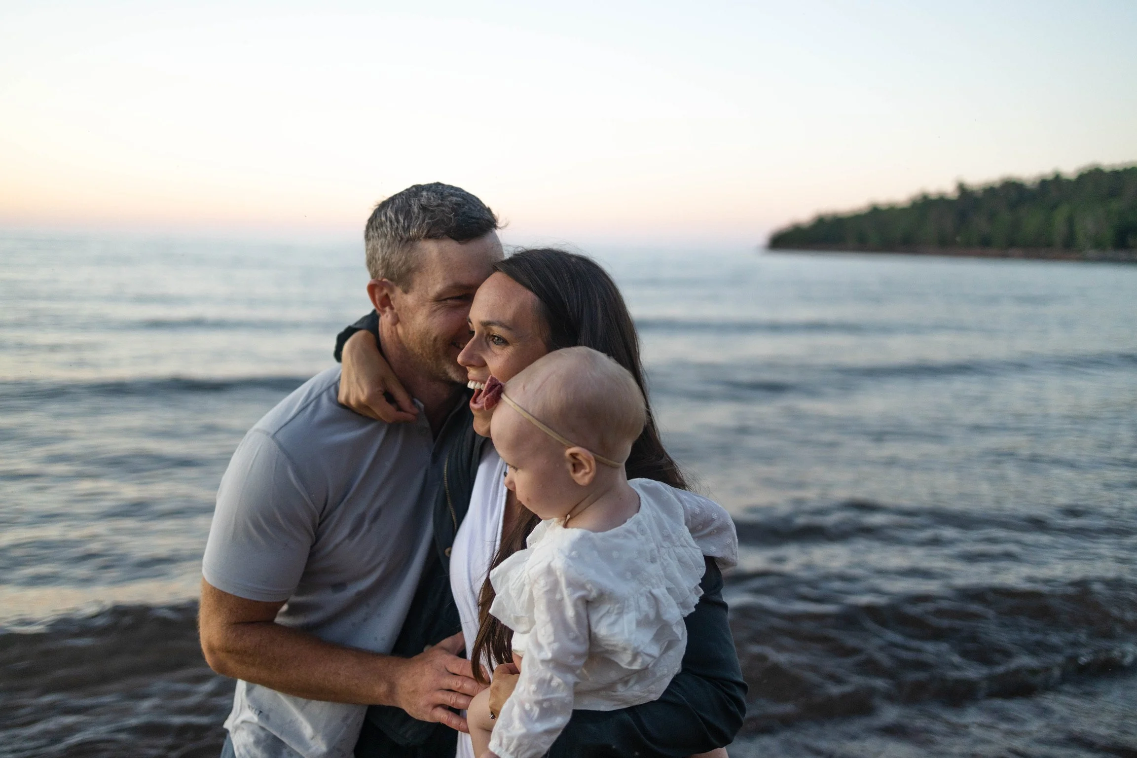 A family of three, a man, a woman, and a young girl, standing on a beach near the ocean at sunset, embracing and smiling.