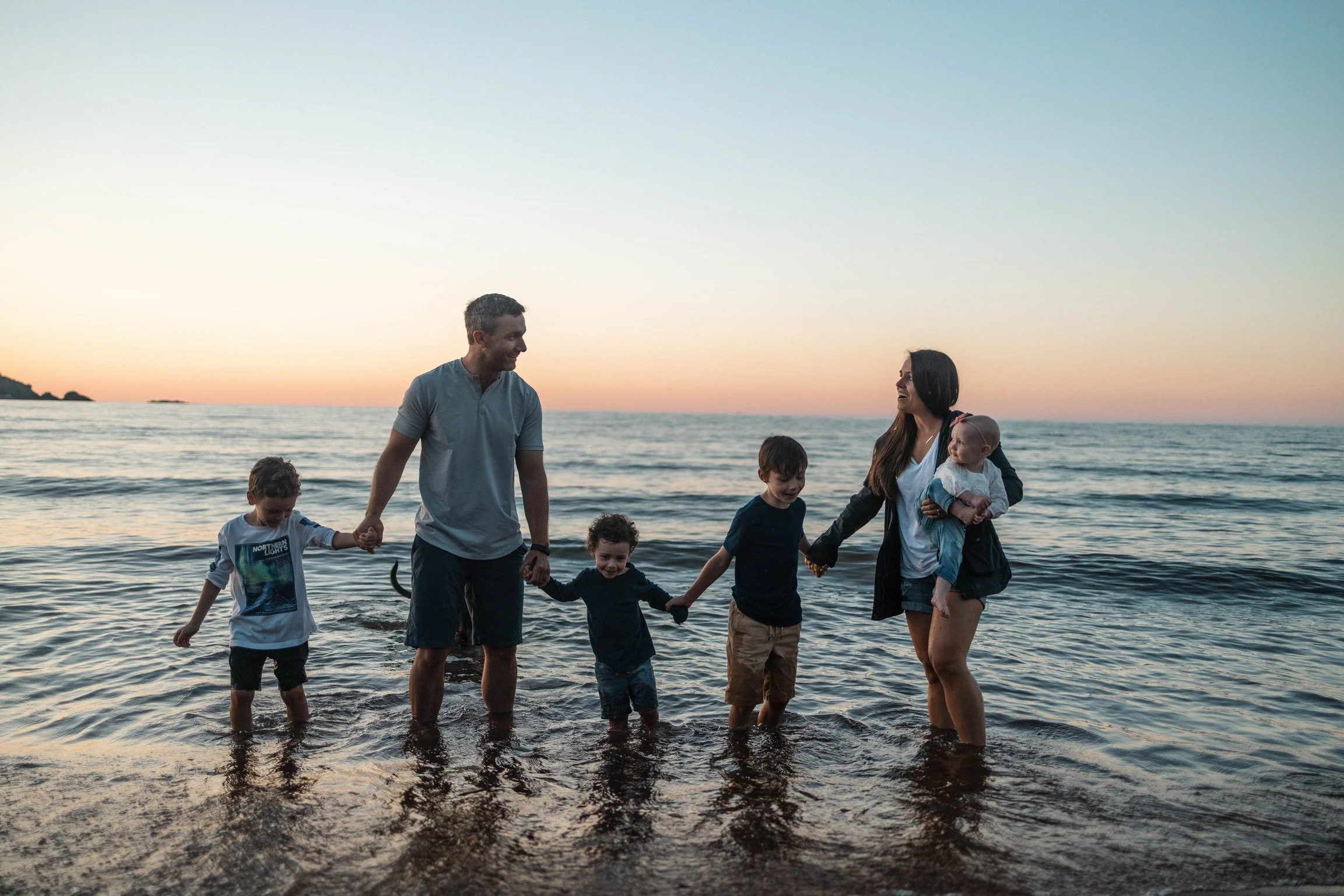 A family of seven, including two adults and five children, holding hands and playing in the shallow water at the beach during sunset.