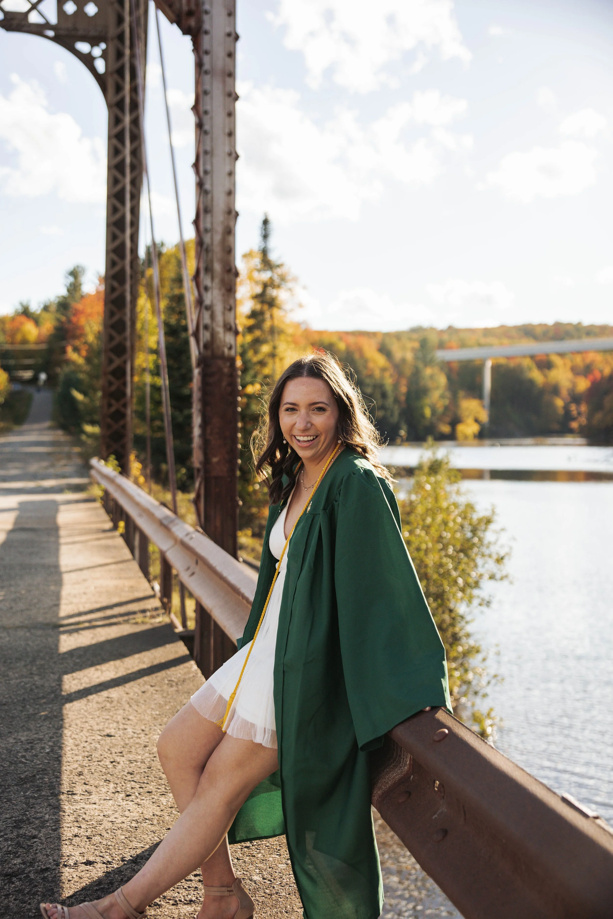 A young woman in a green graduation gown sitting on a bridge railing near a river, smiling at the camera, with autumn trees in the background.