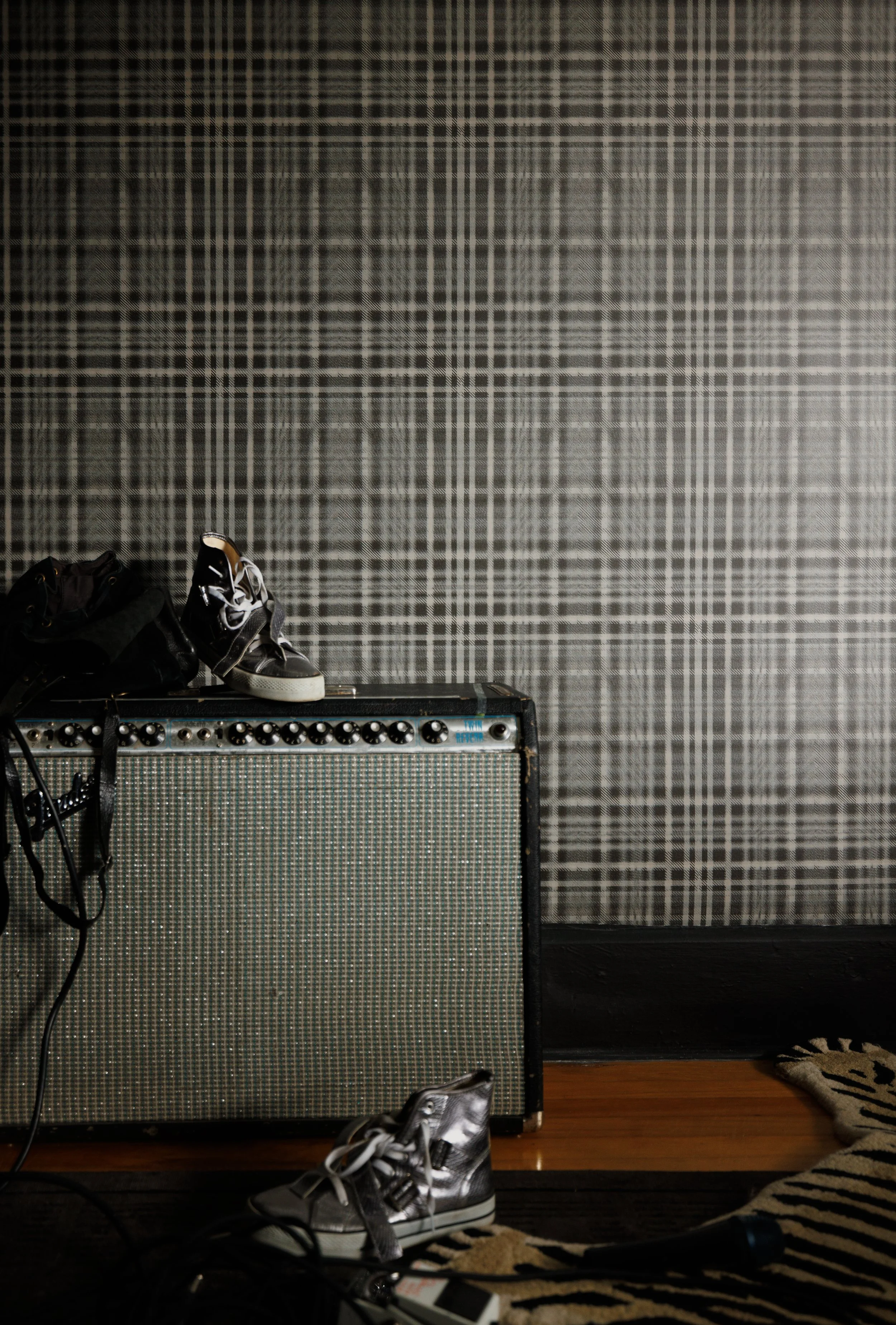 A pair of silver high-top sneakers, a guitar amplifier, a guitar, and some cables are on a wooden floor in front of a dark grey plaid-patterned wall.