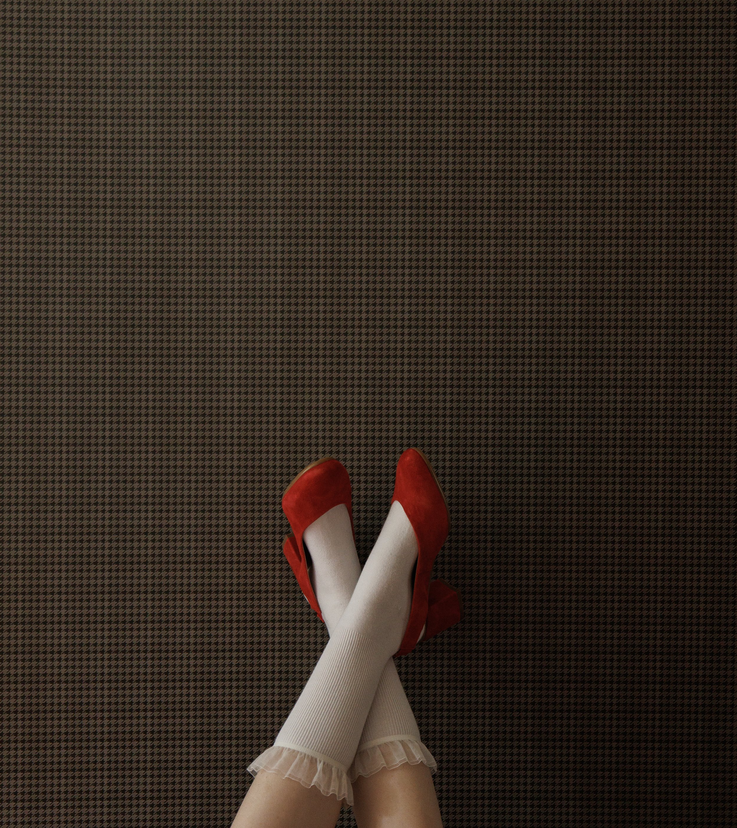 Two feet crossed at the ankles, wearing red high heels and white ruffled socks. The feet are propped up against a wall featuring a dark grey houndstooth wallpaper.