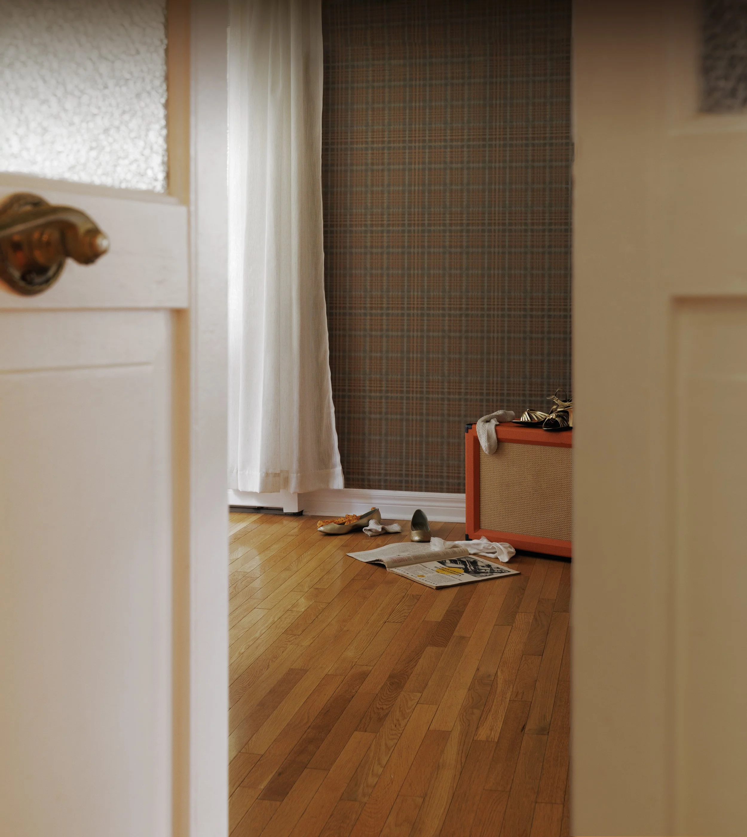 View of a room through a partially open door with a brass doorknob, hardwood flooring, a small orange amp, scattered shoes, and a newspaper on the floor, with a window with closed curtains and a plaid wallpaper on the wall in the background.