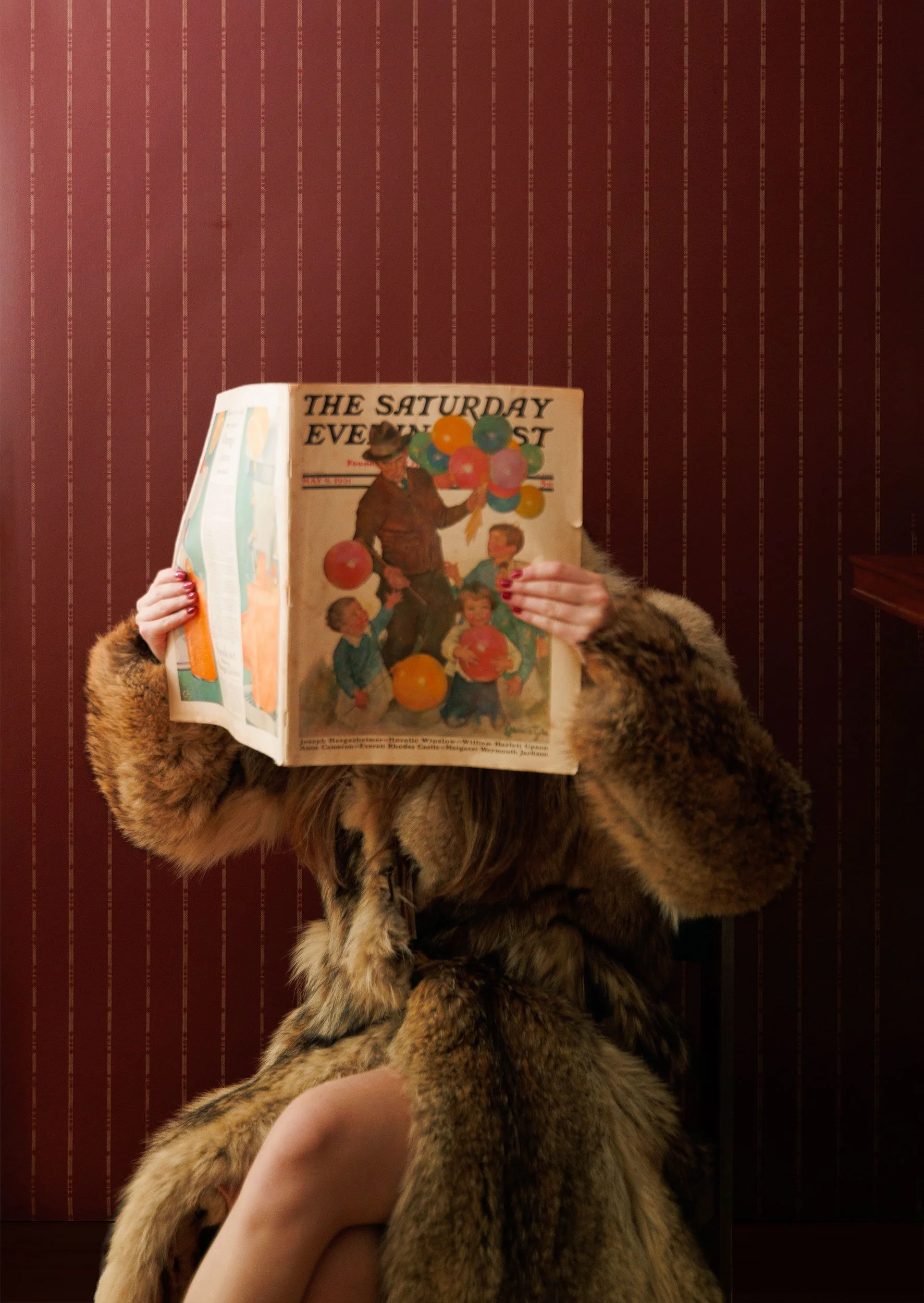 A person holding a vintage magazine titled "The Saturday Evening Post" with an illustration of a man and children playing with balloons. The person is wearing a fur coat, and the background features a deep burgundy wallpaper with beige stripes.