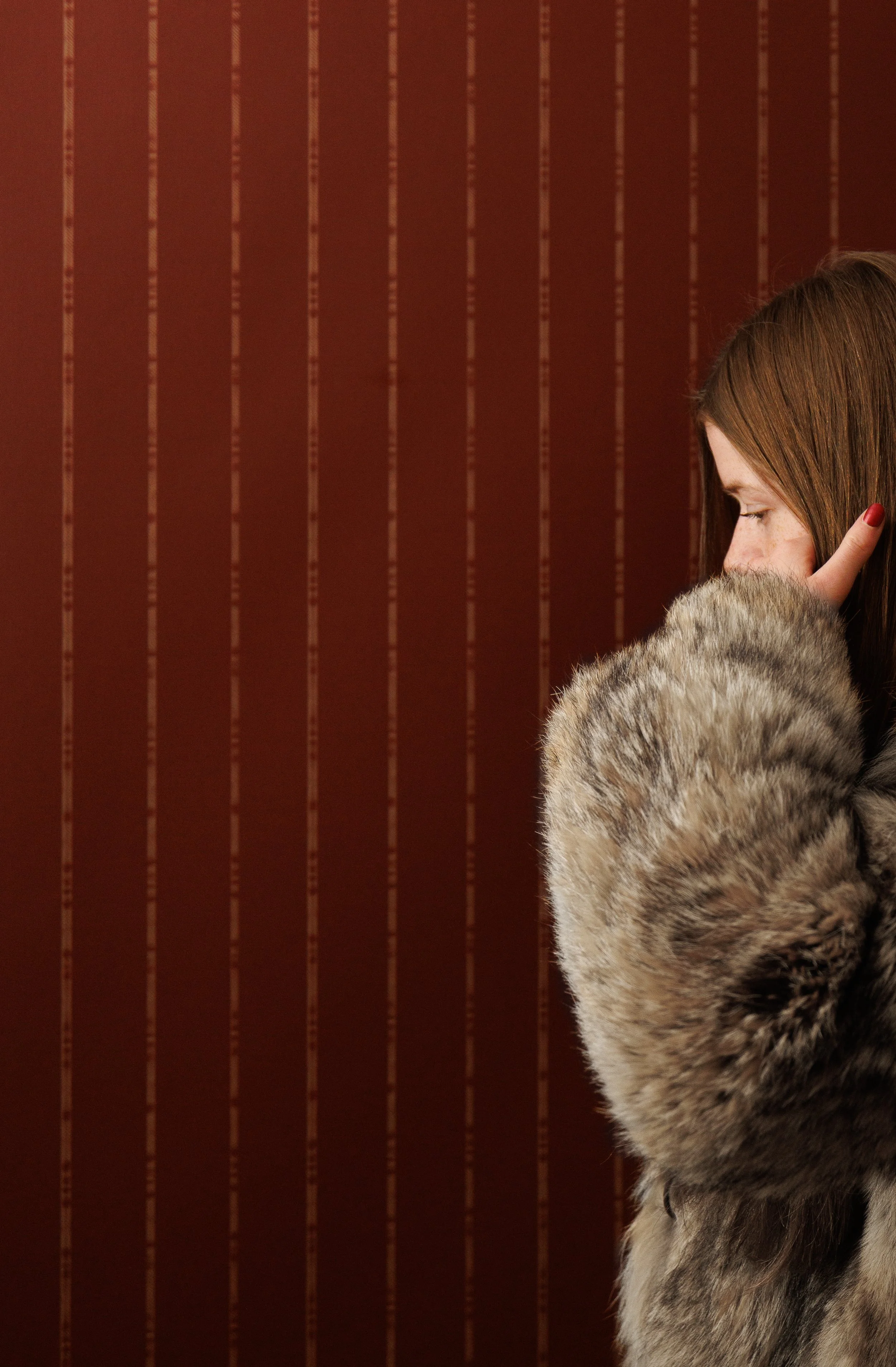 A woman with brown hair and a fur coat, standing sideways with her eyes closed and her hand raised to her face, against a deep burgundy wallpaper with beige vertical lines.