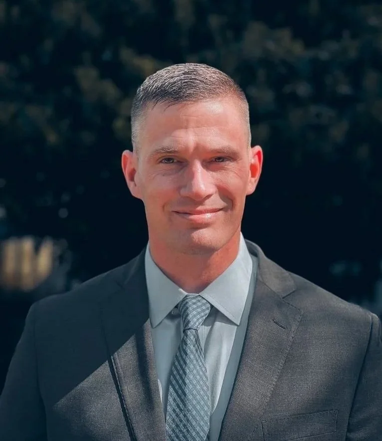 Dr. Marcus R. Van Sickle in a gray suit with a matching tie, smiling outdoors with trees in the background.