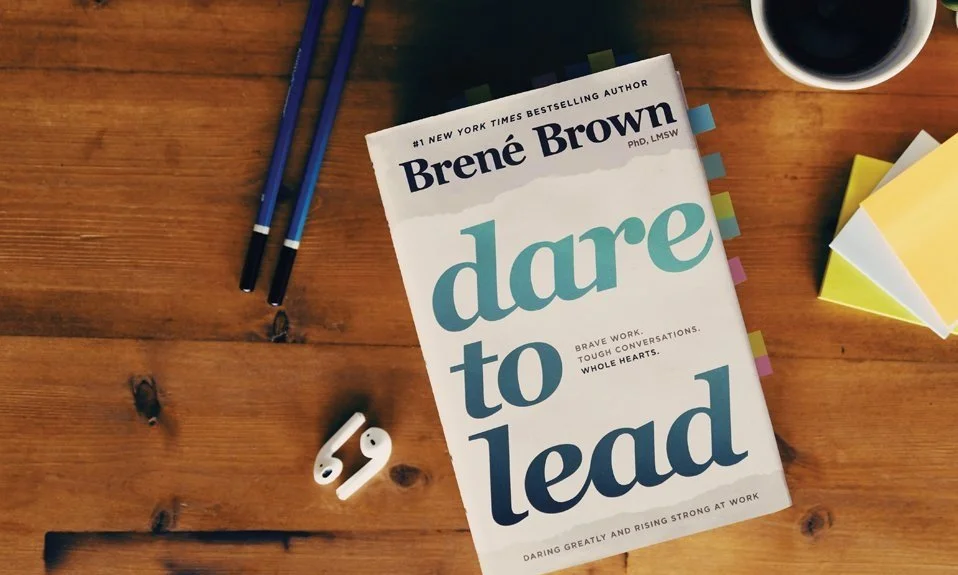 Hardcover copy of Brene Brown's book, Dare to Lead on a wood table with bookmarks, headphones, sticky notes, and pens.