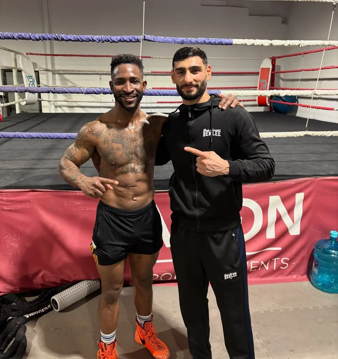 Zwei Männer in einem Boxring, einer nackt mit Tattoos und Sporthose, der andere in Trainingskleidung, beide lächelnd, im Boxstudio.