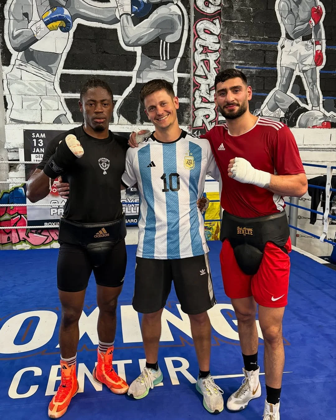 Drei Männer in Boxerausrüstung im Boxring, die sich umarmen. Im Hintergrund eine Wand mit Wandkunst von Boxern. Der mittlere trägt ein argentinisches Trikot, die Männer rechts und links tragen Boxhosen mit Schutzgürteln.