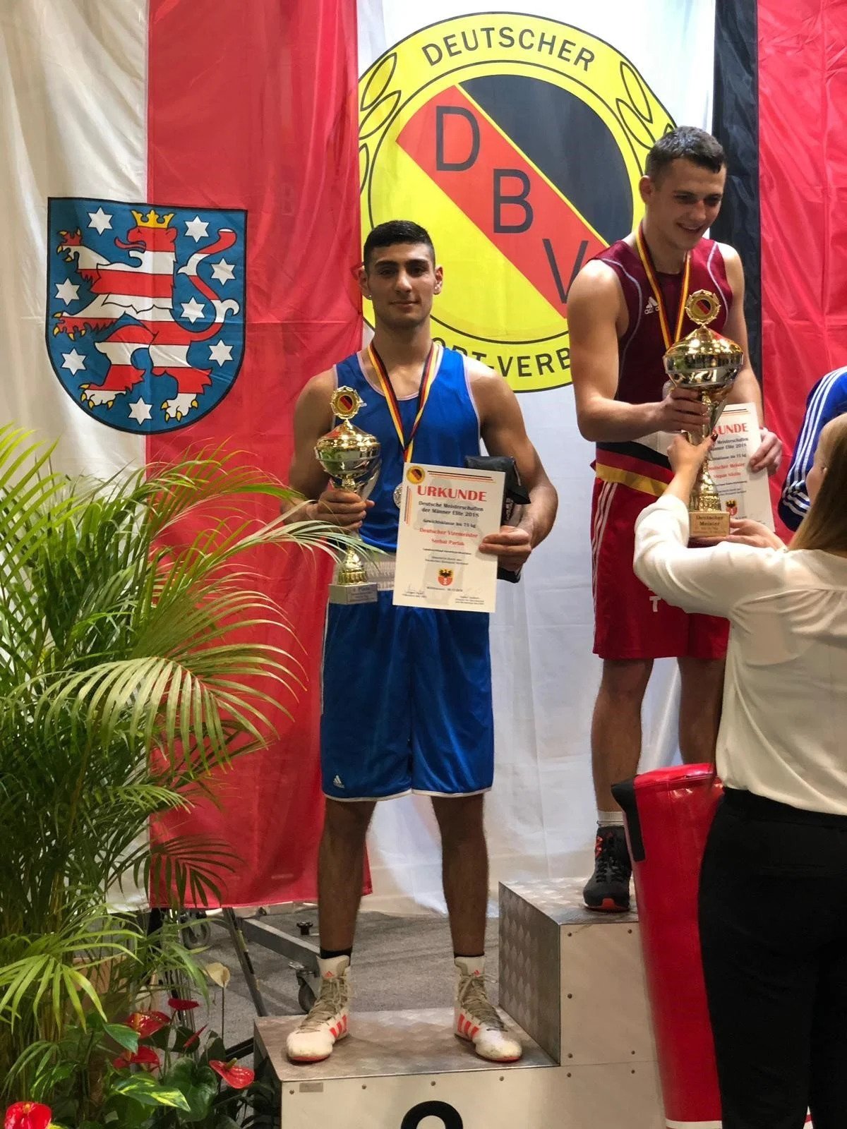 Zwei männliche Boxer auf dem Siegerpodest bei einer Siegerehrung, jeweils mit Trophäen und Urkunden. Hinter ihnen sind Flaggen und das Logo des DBV.