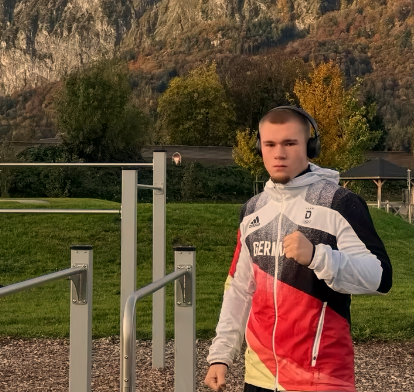 Junge läuft im Freien, trägt Deutschland-Trainingsjacke, Kopfhörer, macht Faustbewegung, im Hintergrund Bäume und Berge.