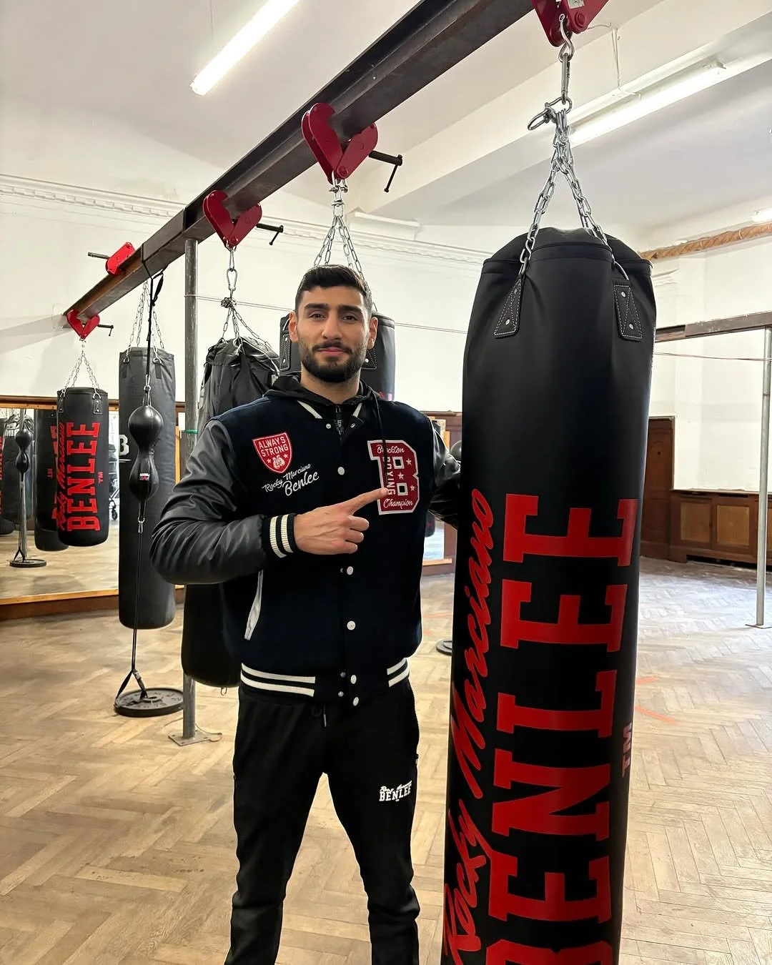 Mann steht in einem Boxstudio neben einem schwarzen Kickbag mit rotem Schriftzug, trägt eine schwarze Jacke mit weißen und roten Details, zeigt auf die Tasche.