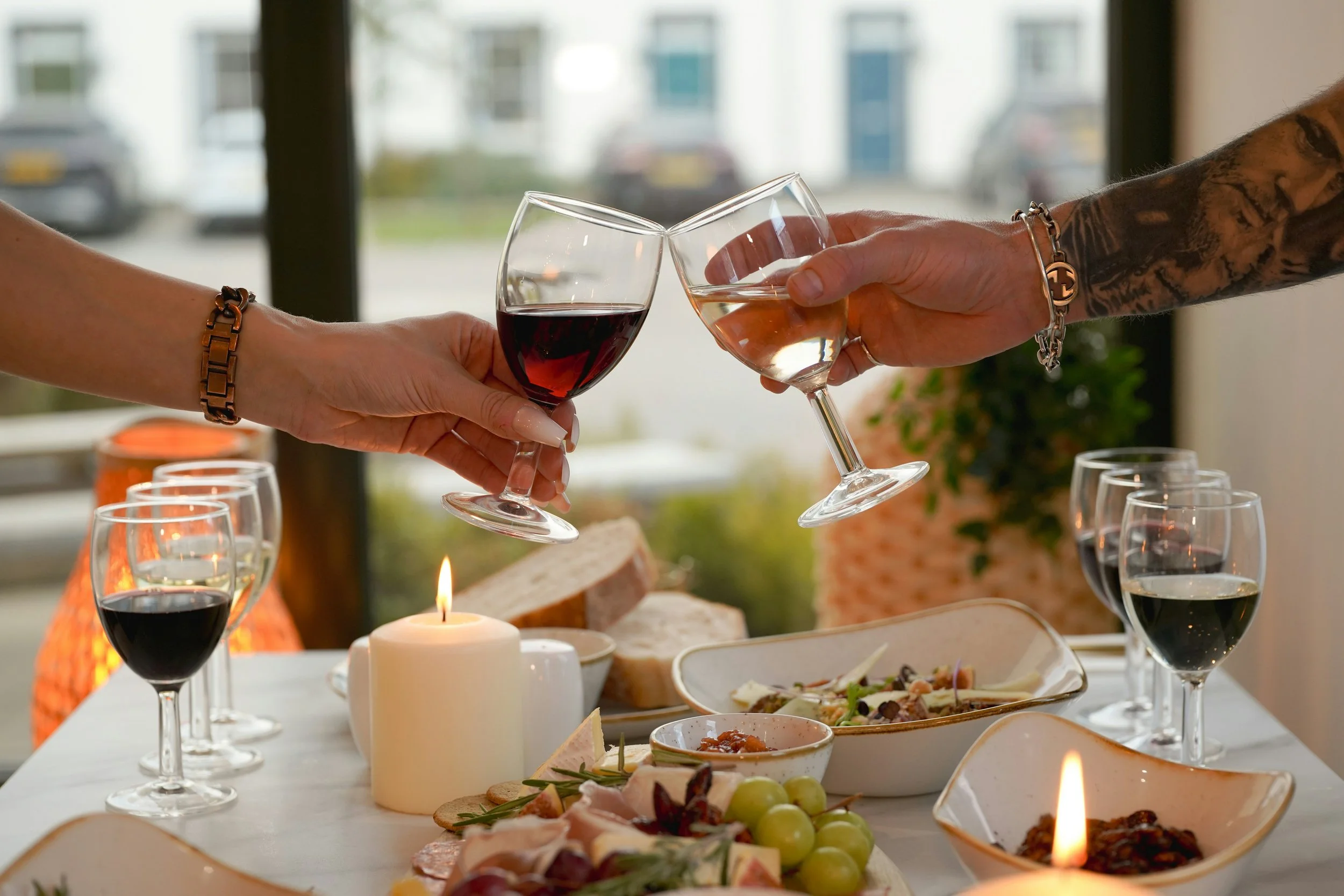 A close-up image of two hands toasting with glasses of red and white wine, surrounded by various dishes, a lit candle, and other wine glasses, creating an inviting dining atmosphere.