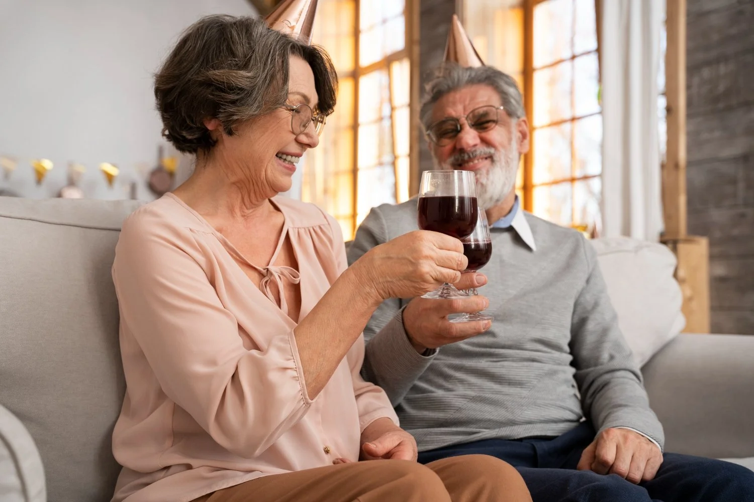 A woman wearing a light pink blouse and glasses smiles as she raises a glass of dark liquid alongside a bearded man in a grey sweater, both wearing festive party hats.