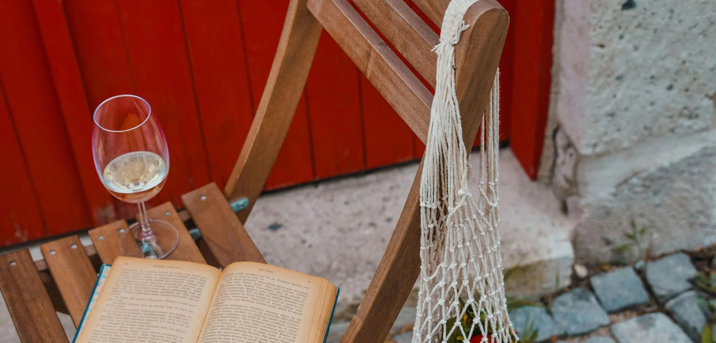 A glass of non-alcoholic white wine and an open book rest on a wooden folding chair, with a cream-colored macrame string bag hanging from the side near a red door.